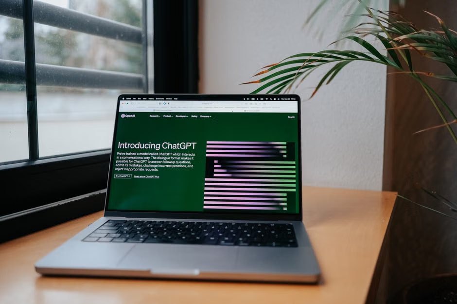 A laptop screen displays the ChatGPT interface, positioned on a desk next to a window, suggesting a modern home office environment.