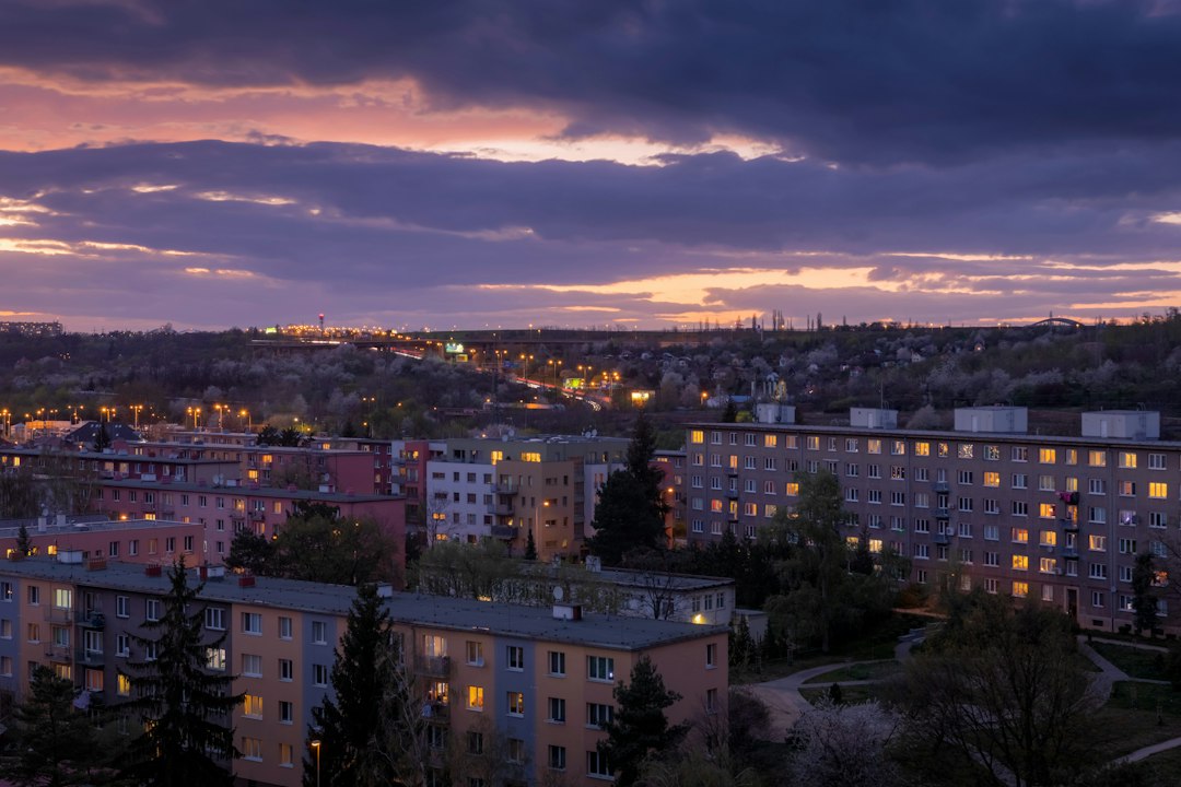 Muž v obleku s bezpečnostní přilbou na hlavě si prohlíží stavební dokumenty na pozadí oploceného staveniště u bytového domu.