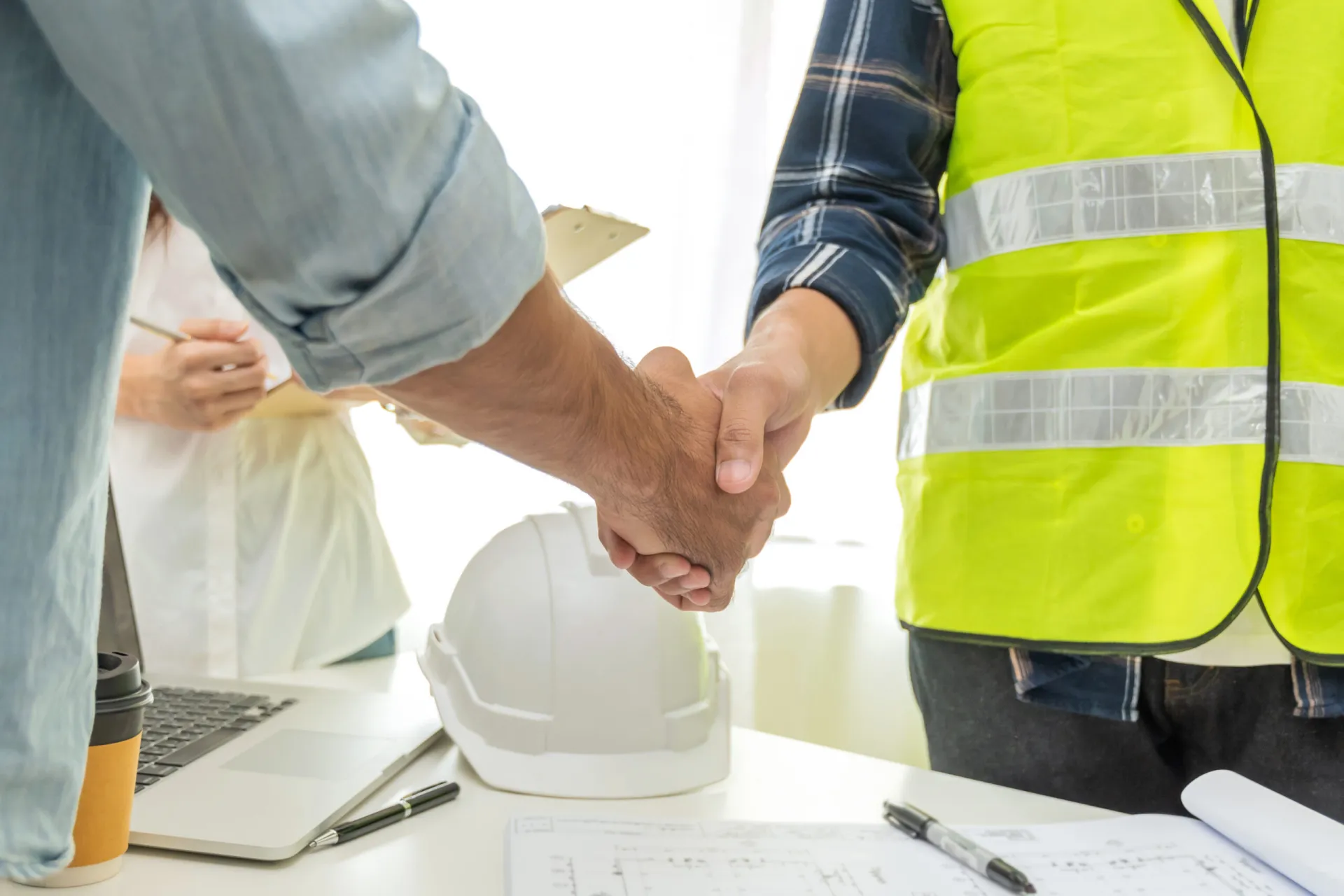 Two professionals in business attire shaking hands in front of a modern building under construction, with scaffolding and cranes visible in the background.