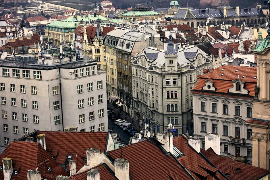 A high-angle view of Prague's historic district, showing a dense collection of buildings with red-tiled roofs, spires, and intricate architectural details under a clear sky.