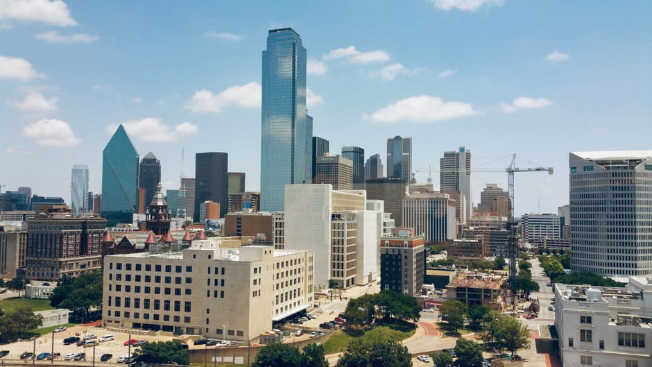 An aerial view of the Dallas skyline, showing numerous modern skyscrapers against a clear blue sky.